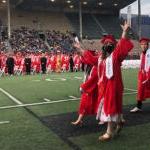 Four graduates in cap and gown walk across a sports field